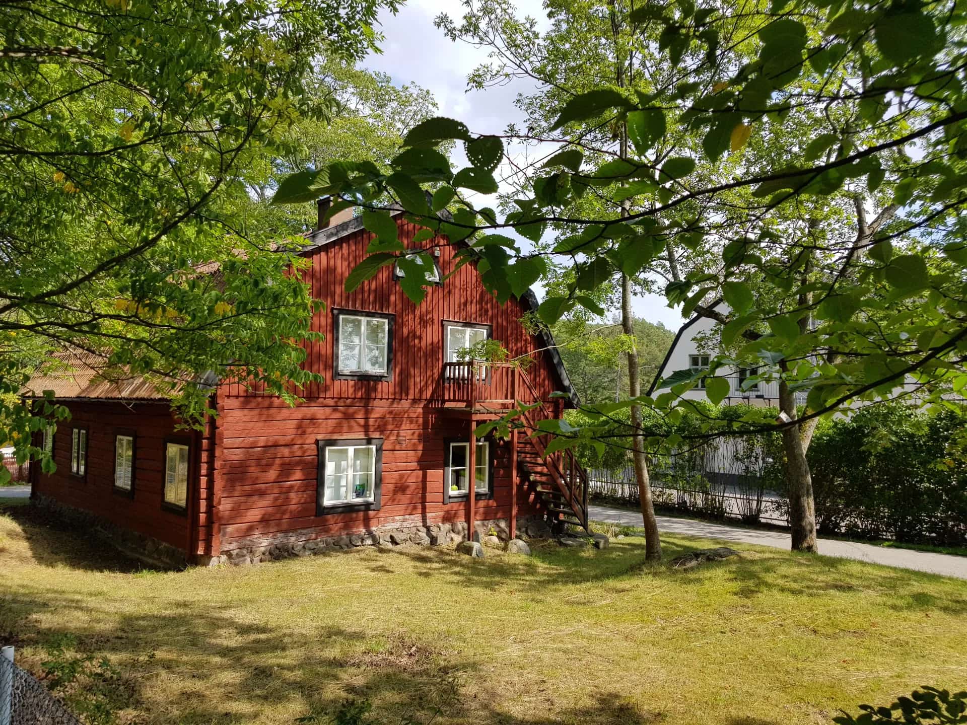 Red wooden house with white windows and green lawn, partially obscured by trees
