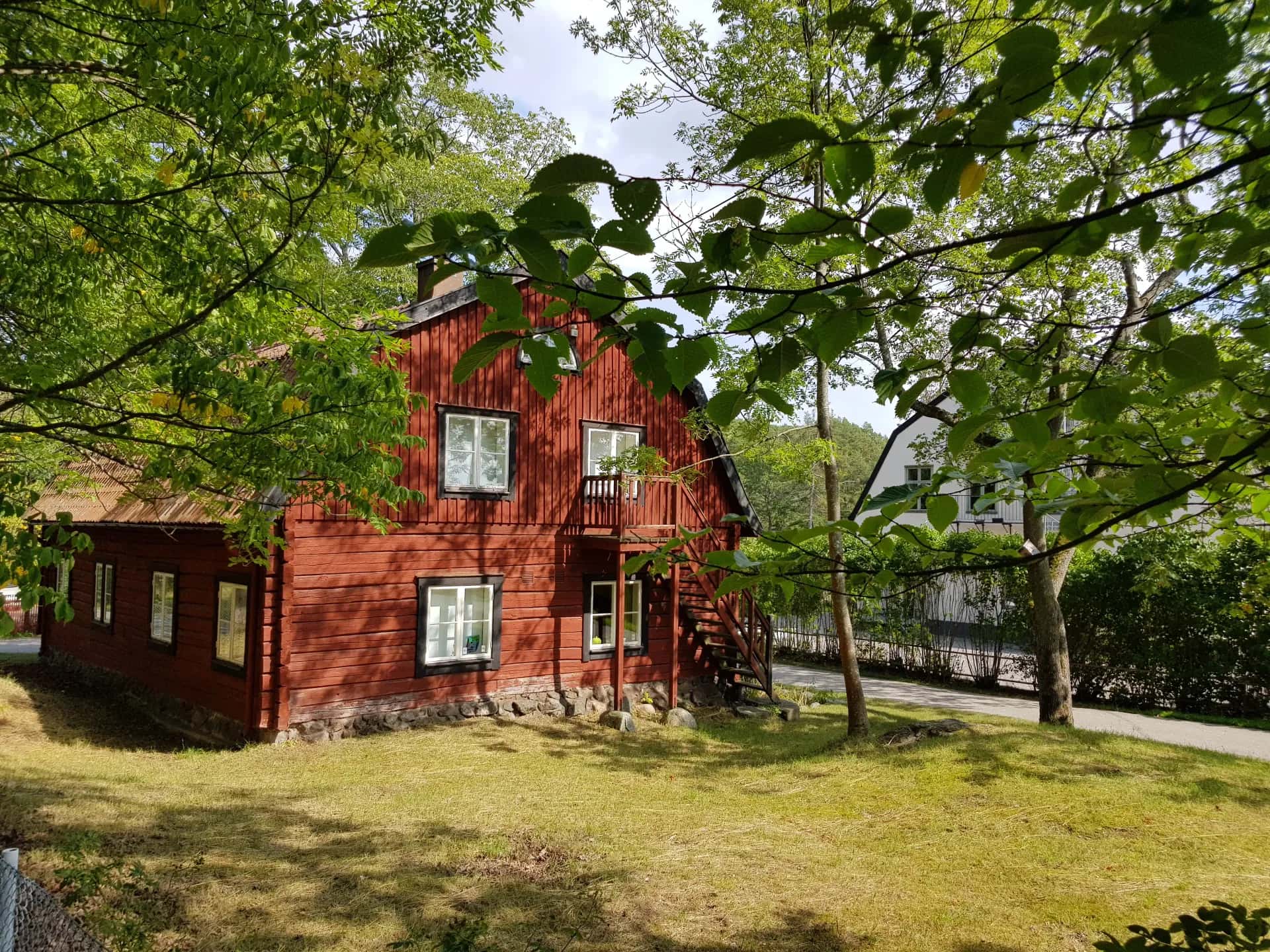 Rött trähus med vita fönster och trappa, omgivet av gröna träd och gräsmatta