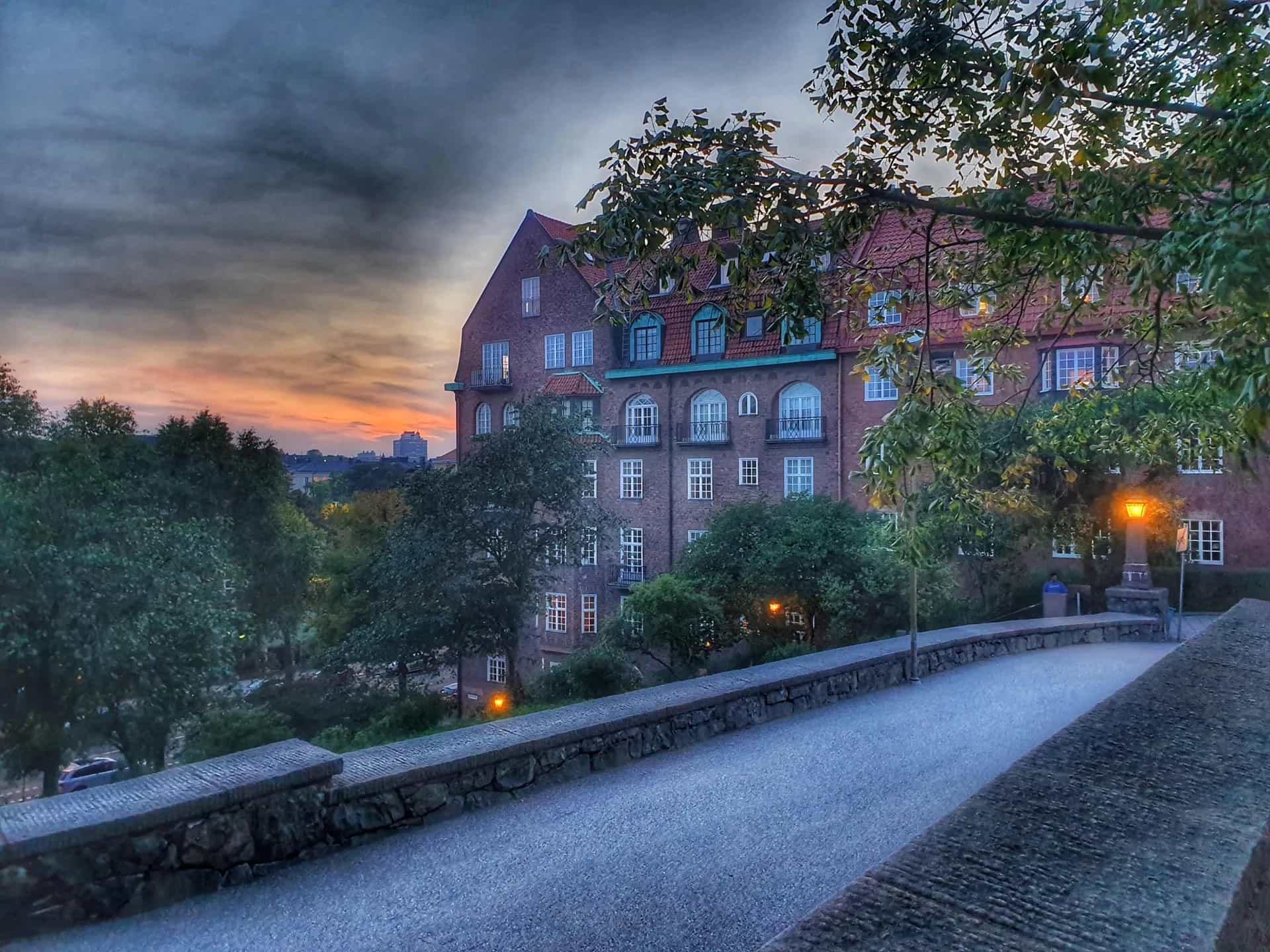 Gångväg med stenmur, rött tegelhus och träd under grå himmel med solnedgång