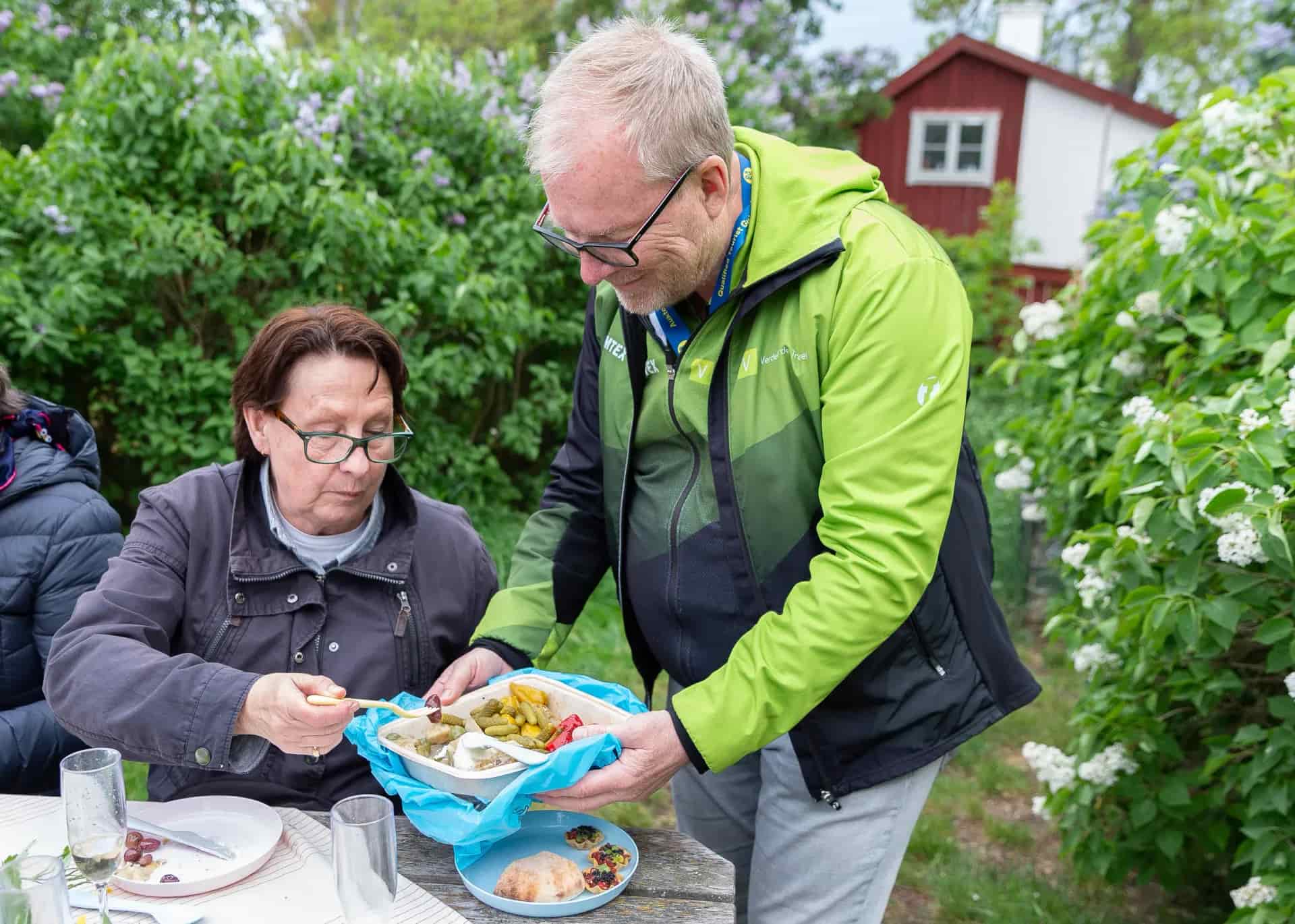 Two people outdoors at a table with food, one of whom is pouring food. Bushes and red building in the background