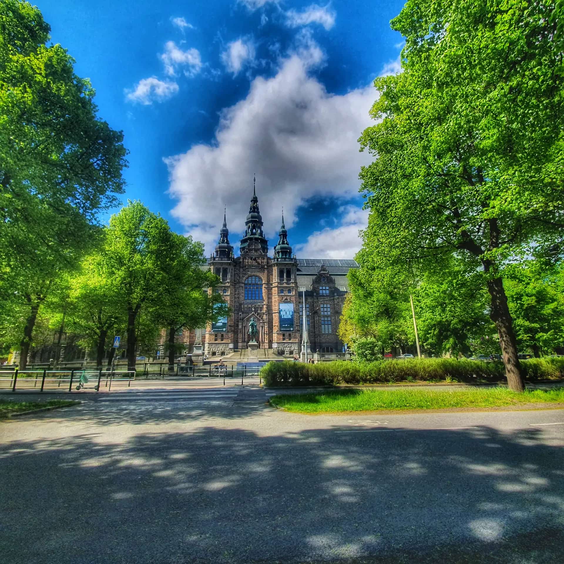 A brown building with a tower seen through green trees, under a blue sky with clouds