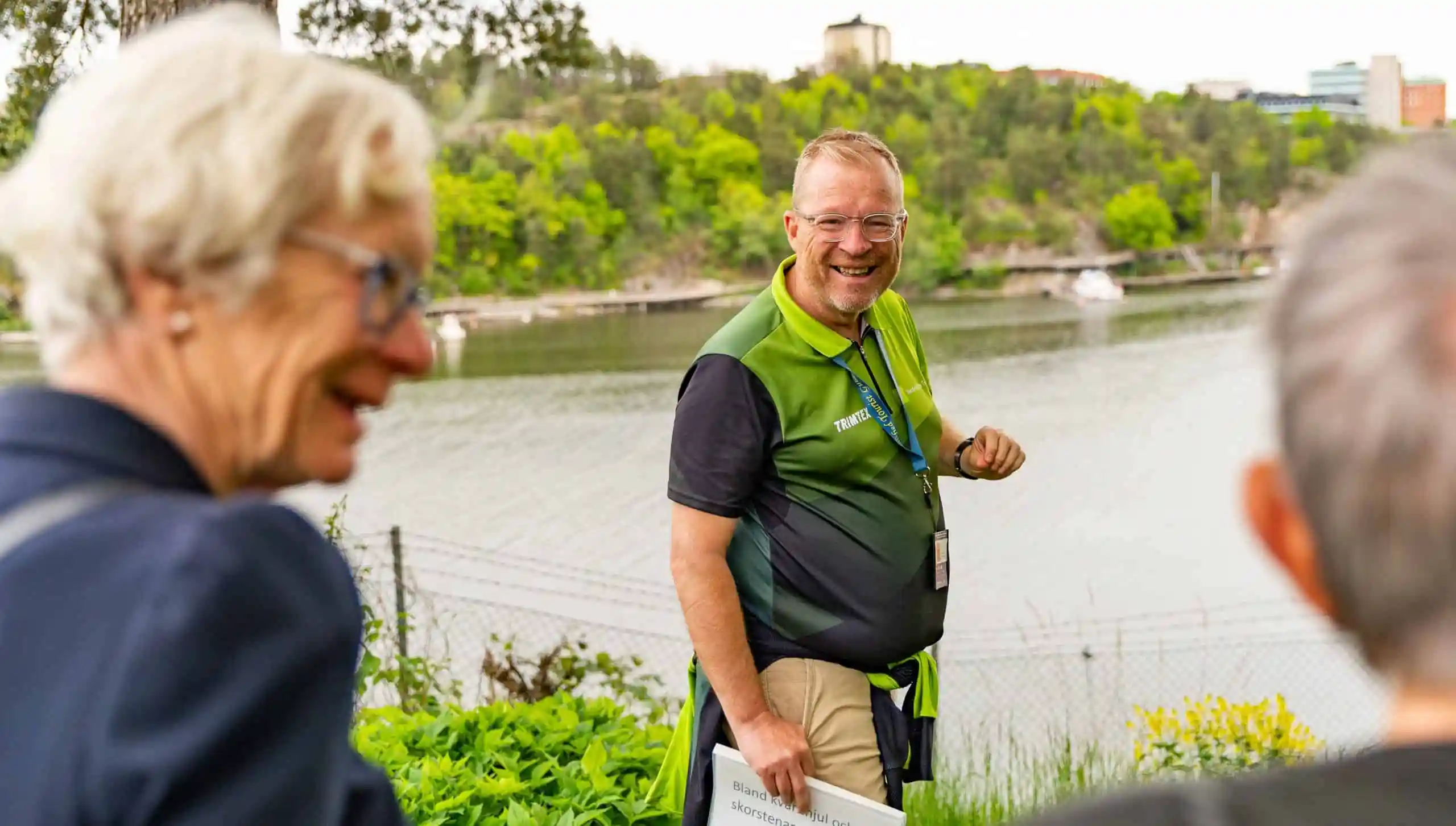 Man in green shirt with text stands by water and smiles at camera. Women in foreground